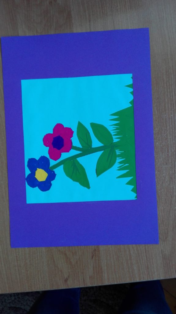 Na zdjęciu jest praca pokazująca dwa kwiaty wyrastające z jednej łodygi. Jeden jest różowy, drugi fioletowy. Praca wyklejana z kolorowego papieru.